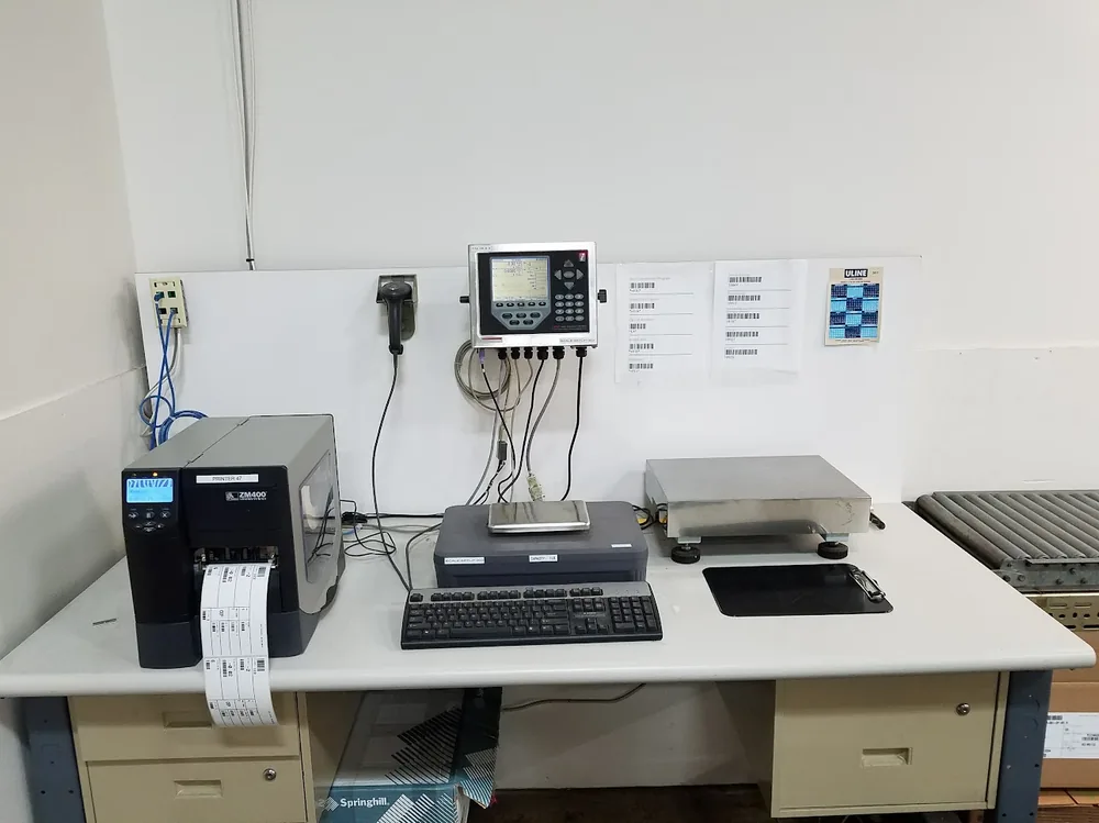 Counting and labeling workstation: a Zebra ZM400 printing labels beside a bench scale platform and wall-mounted indicator, with barcode samples posted above.