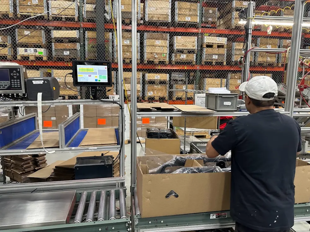 An operator boxing parts at a conveyor-side packing station, with a touchscreen terminal, scale indicator and Zebra label printer mounted at the line and pallet racking behind.
